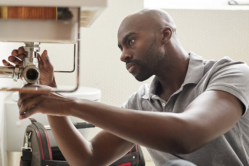 5 Summer Plumbing Tips to Avoid Water Waste in Greater Houston. Photo of a plumber inspecting under a bathroom or kitchen sink.